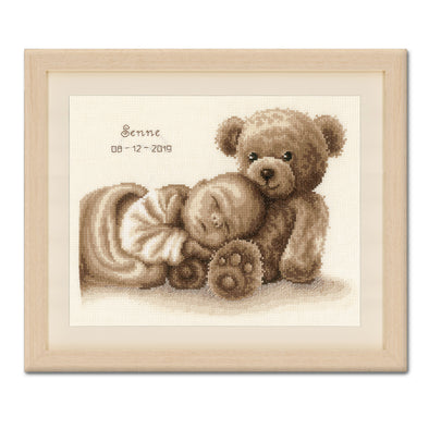 Framed cross-stitched picture of a teddy bear holding a baby bootie on a white background